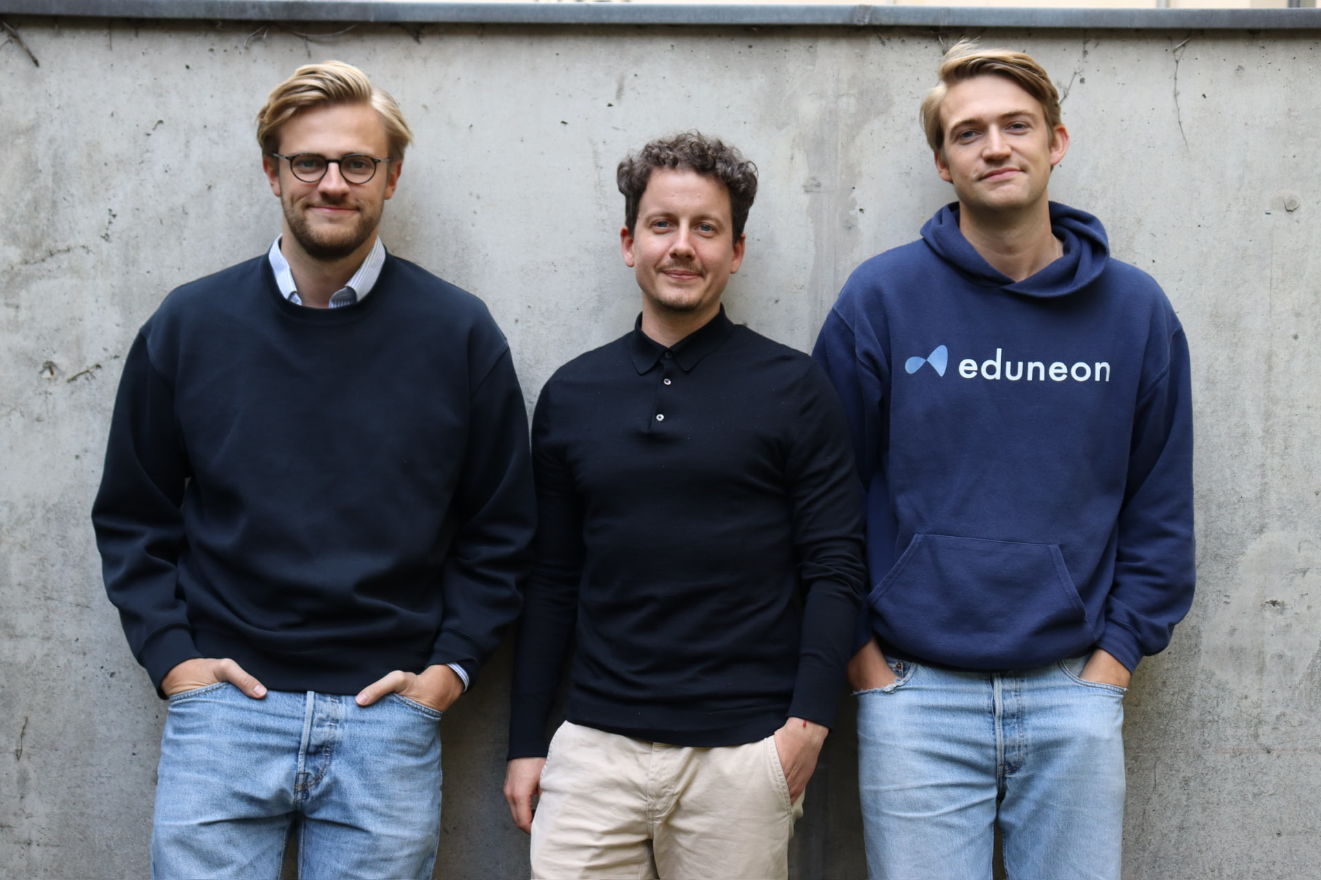 Drei Eduneon-Mitarbeitende, einer im Kapuzenpullover mit Eduneon-Schriftzug, vor einer Betonwand